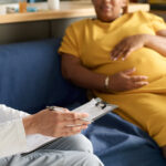 Pregnant woman in a yellow dress sitting on a couch during a consultation with a healthcare provider holding a clipboard and pen.