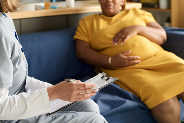 Pregnant woman in a yellow dress sitting on a couch during a consultation with a healthcare provider holding a clipboard and pen.