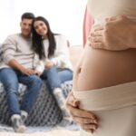 A pregnant woman holding her belly while an intended couple smiles and watches from the couch nearby.