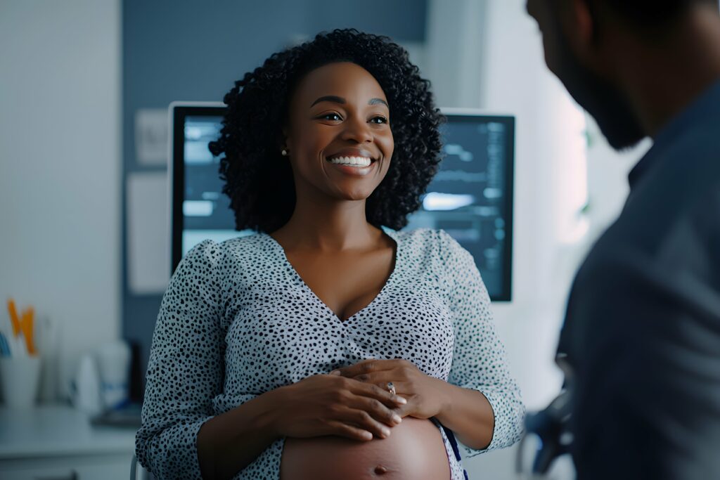 Pregnant woman smiling during a prenatal appointment, seated in a medical setting with an ultrasound screen in the background.