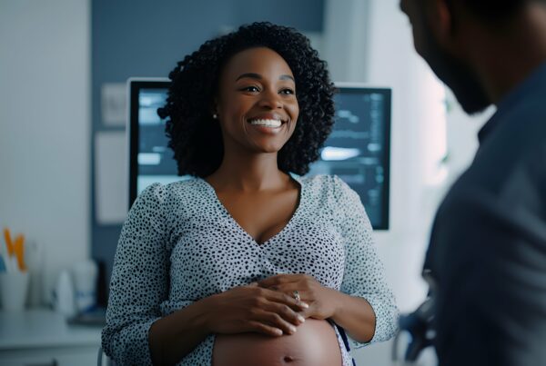Pregnant woman smiling during a prenatal appointment, seated in a medical setting with an ultrasound screen in the background.