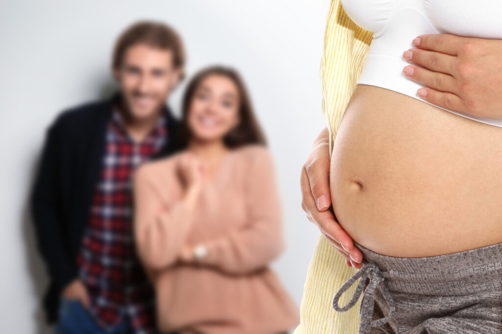 A pregnant surrogate mother gently cradles her bare belly in the foreground, while the intended parents smile in the blurred background, standing close together.