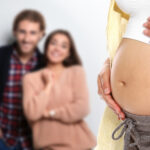 A pregnant surrogate mother gently cradles her bare belly in the foreground, while the intended parents smile in the blurred background, standing close together.