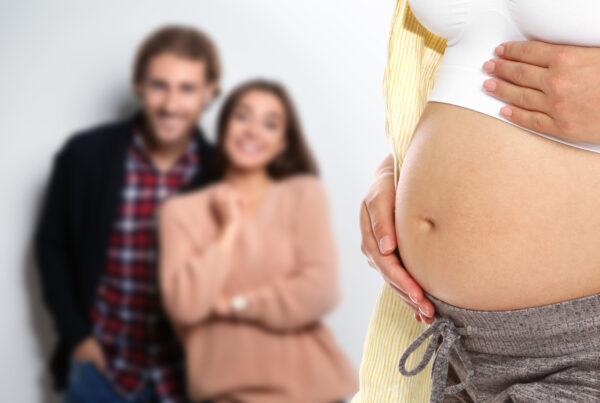 A pregnant surrogate mother gently cradles her bare belly in the foreground, while the intended parents smile in the blurred background, standing close together.