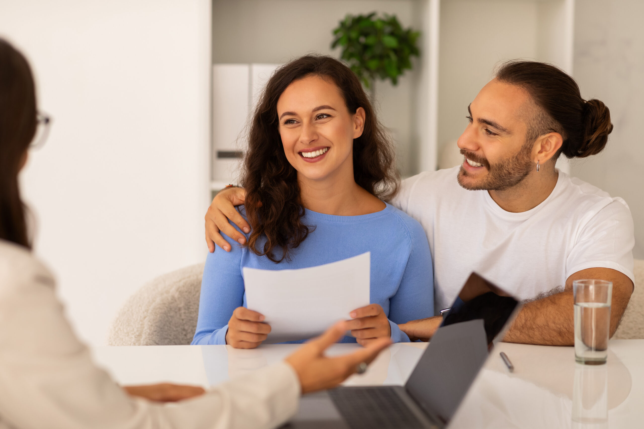 Two smiling intended parents smile while sitting at a desk across from a professional who is assisting them with the surrogacy process. The woman holds paperwork while the man wraps his arm around her.
