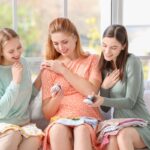 Three women smiling together on a couch during a baby shower, holding baby clothes and shoes. The central woman is pregnant and wearing an orange polka-dot dress, surrounded by supportive friends.