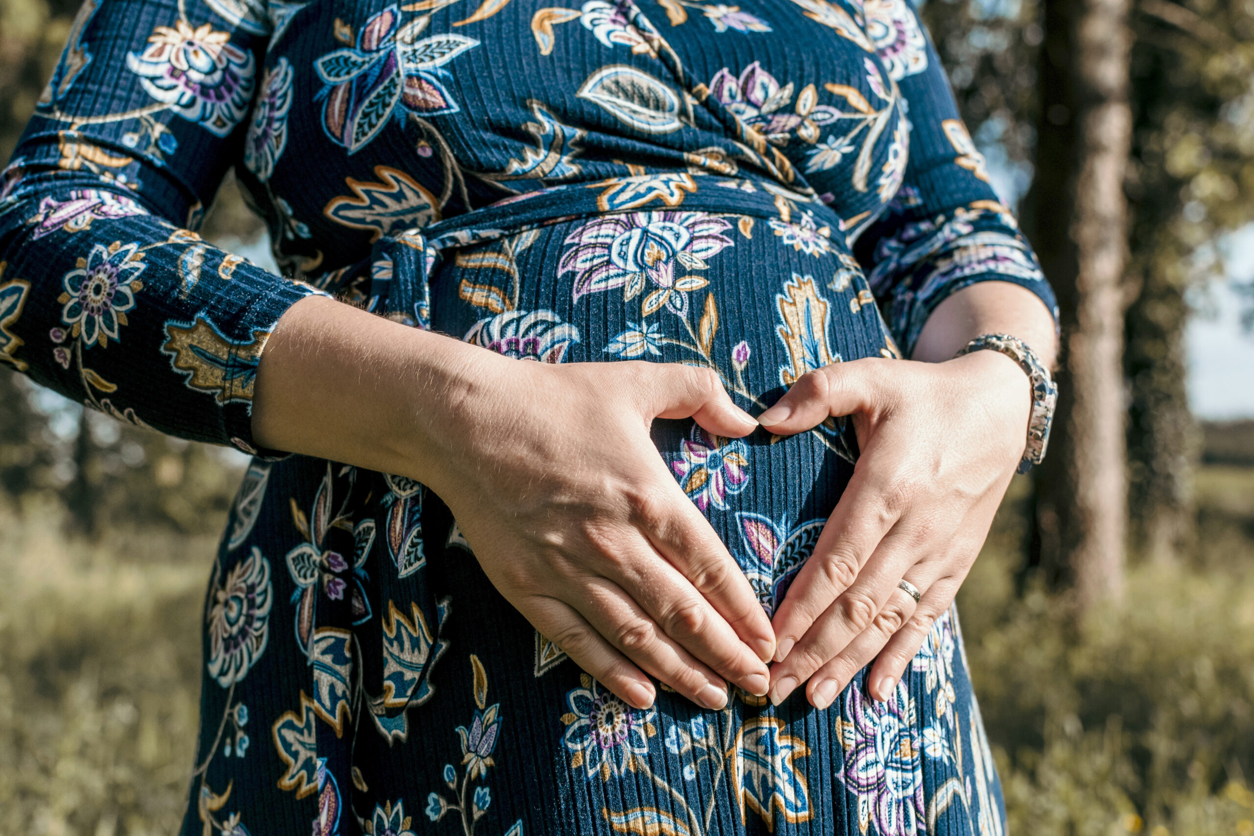 Close-up of a pregnant woman standing outdoors in a blue floral dress, forming a heart shape with her hands over her baby bump to symbolize love and connection during pregnancy.