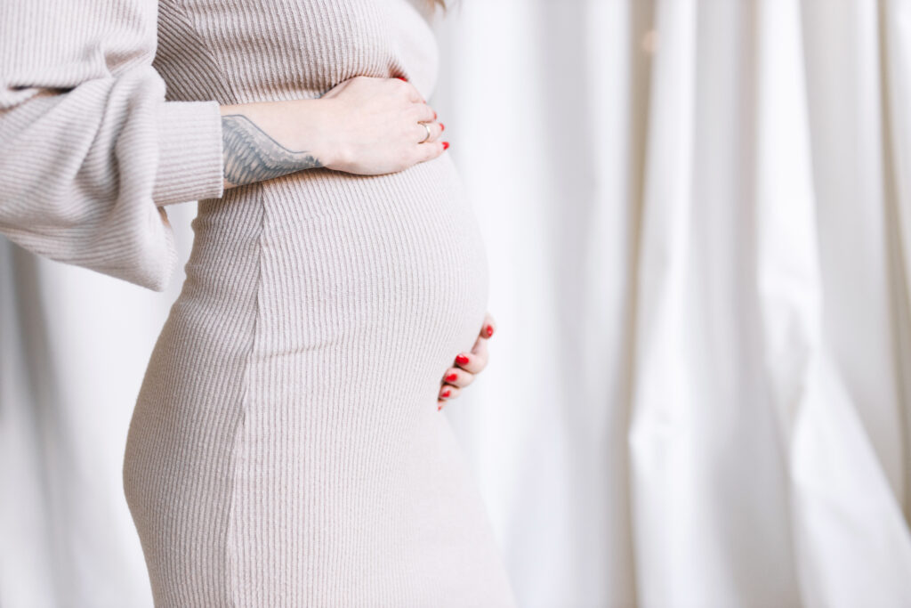 Close-up of a pregnant woman wearing a beige ribbed dress, gently cradling her belly with both hands. Her red nails and a tattoo on her forearm are visible.