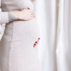 Close-up of a pregnant woman wearing a beige ribbed dress, gently cradling her belly with both hands. Her red nails and a tattoo on her forearm are visible.