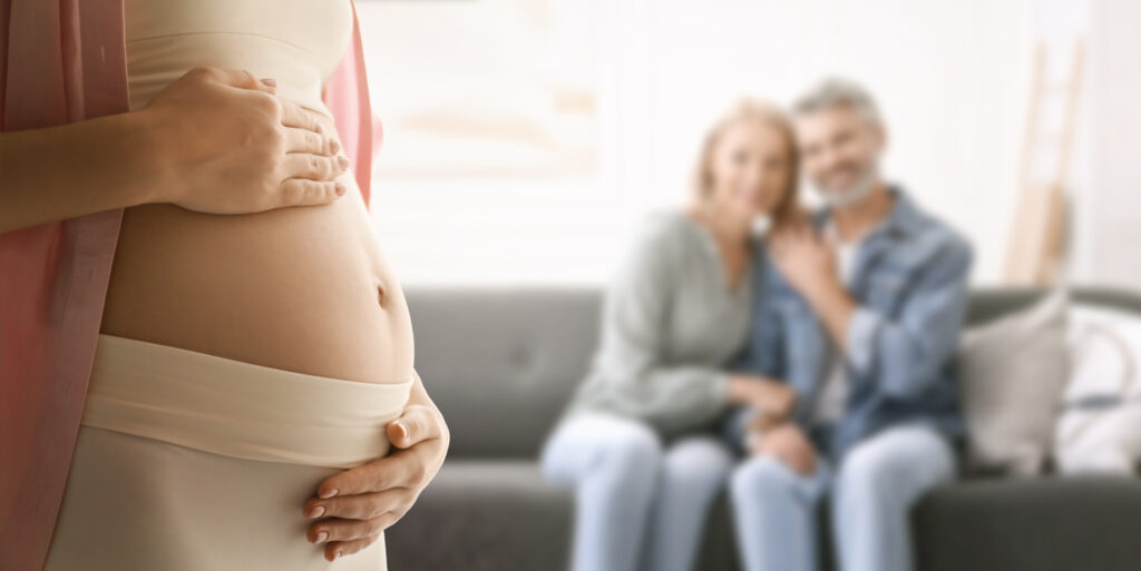 A close-up of a pregnant woman gently holding her belly, with two intended parents sitting together in the background, smiling softly.