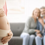 A close-up of a pregnant woman gently holding her belly, with two intended parents sitting together in the background, smiling softly.