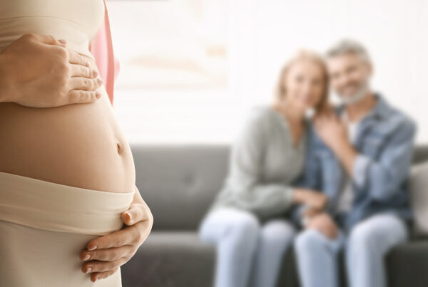 A close-up of a pregnant woman gently holding her belly, with two intended parents sitting together in the background, smiling softly.