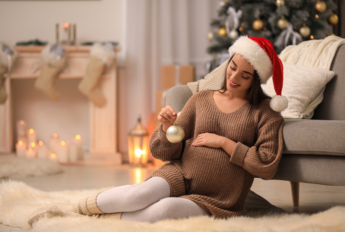 Smiling pregnant woman in a Santa hat sitting on the floor, holding a holiday ornament near her belly, with a Christmas tree and stockings in the background.