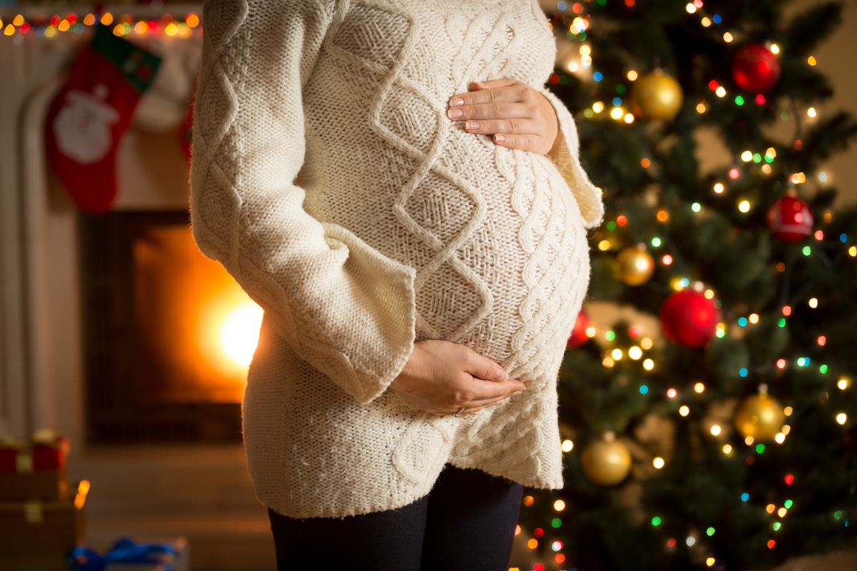 Pregnant woman in a cream sweater cradling her belly in front of a decorated Christmas tree and fireplace.