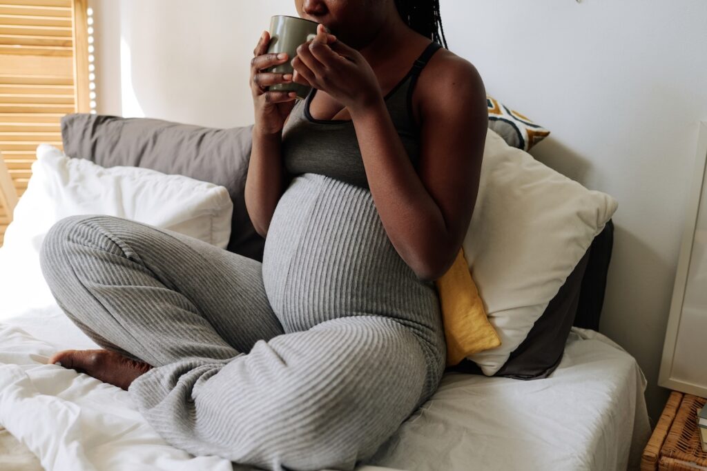 Pregnant woman sitting on a bed in comfortable loungewear, holding a mug and relaxing with supportive pillows around her.