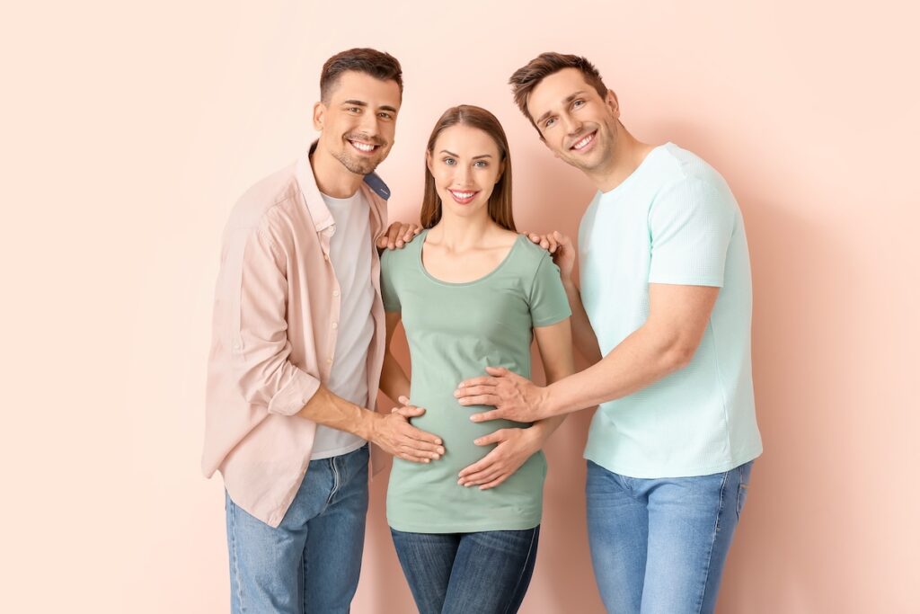 Two men stand on either side of a smiling pregnant woman, each resting a hand on her shoulder or belly against a soft peach background.