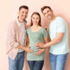 Two men stand on either side of a smiling pregnant woman, each resting a hand on her shoulder or belly against a soft peach background.