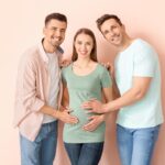 Two men stand on either side of a smiling pregnant woman, each resting a hand on her shoulder or belly against a soft peach background.