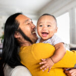 Smiling father holding a happy baby in a bright home, showing a warm parent-child moment.
